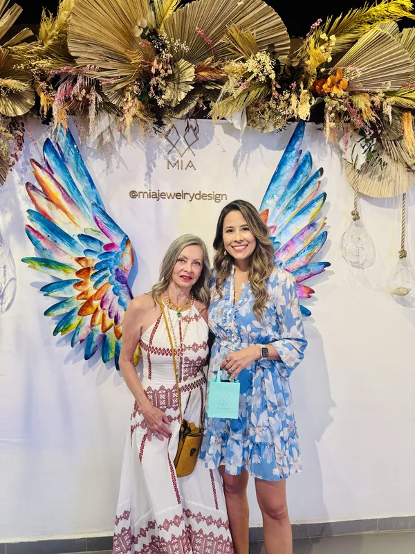 Dos invitadas con vestidos blanco y azul sostienen una bolsa de regalo aqua de MIA, enmarcadas por las alas de ángel pintadas en un evento MIA