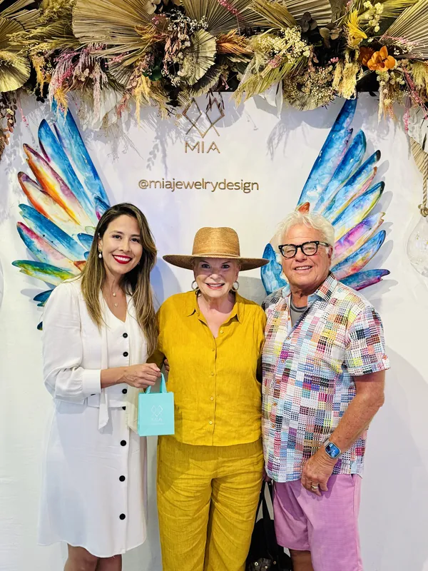 Imelda de blanco con dos coleccionistas bajo un arco de flores secas; la clienta de amarillo sostiene su bolsa de regalo aqua de MIA