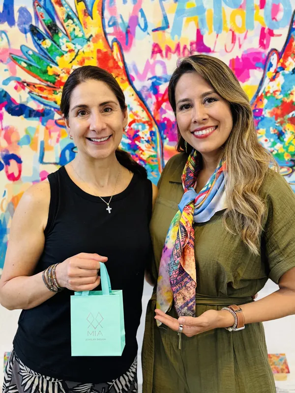 Dos amigas ante el mural colorido de alas de ángel: una sostiene la bolsa aqua de MIA y la otra lleva su collar de cruz de plata